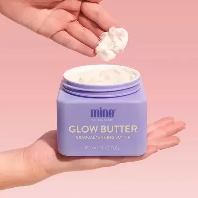 Frasco lilás de creme com texto GLOW BUTTER, mãos aplicando creme, fundo rosa.