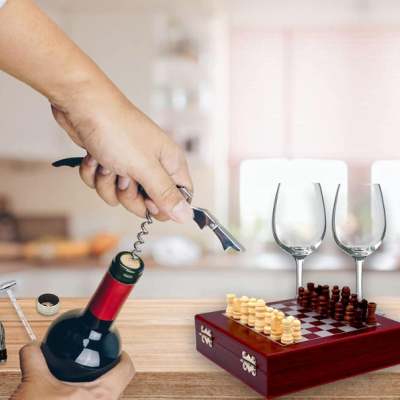 Mão a abrir garrafa de vinho com saca-rolhas, taças e tabuleiro de xadrez