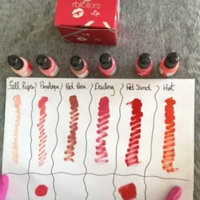 Seis frascos de verniz para unhas com cores diferentes sobre papel com amostras e nomes das cores, com caixa vermelha ao lado.