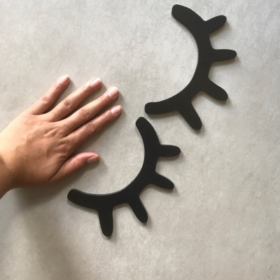 Duas peças decorativas pretas em forma de pestanas sobre superfície cinzenta com mão à esquerda