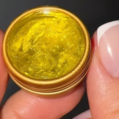 frasco pequeno amarelo com gel segurado por mão com unha branca sobre fundo preto
