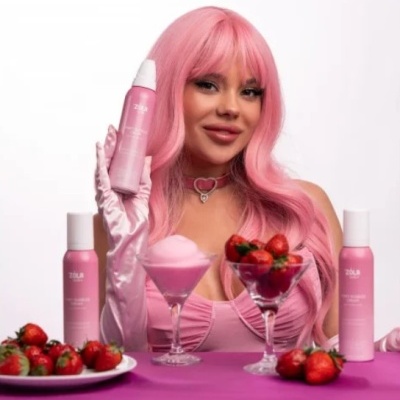 Mulher com cabelo cor-de-rosa segura produto ZOLA rodeada de morangos e taças sobre mesa cor-de-rosa