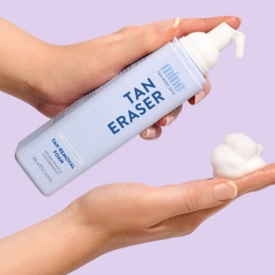 Frasco azul claro de espuma para remoção de bronzeado sendo aplicado pelas mãos sobre fundo lilás.