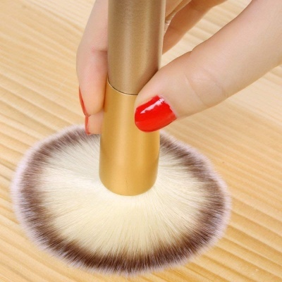 Pincel de maquilhagem com cerdas esbranquiçadas e cabo dourado sobre madeira clara.