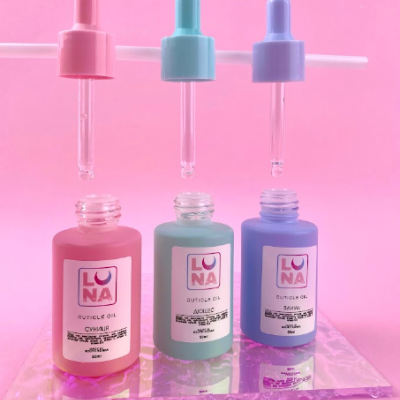 Três frascos de óleo para cutículas de cores rosa, azul e lilás com tampas conta-gotas, sobre fundo rosa.