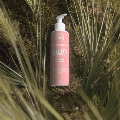 Frasco rosa do produto purify mousse da Cacau Chic em meio a plantas secas e musgo