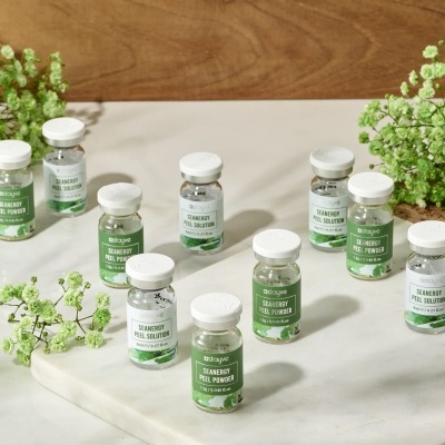 Frascos pequenos de vidro com tampas brancas e rótulos verdes sobre superfícies branca e madeira, com flores verdes ao redor.