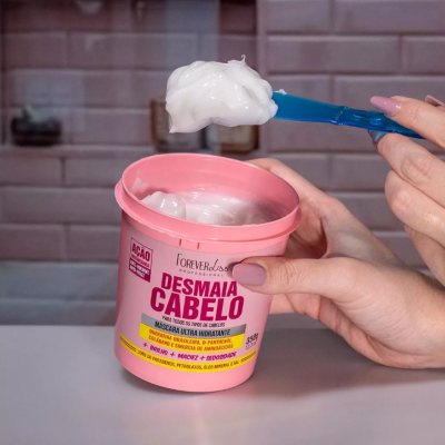 Pote cor-de-rosa com máscara capilar branca e espátula azul segurando produto cremoso