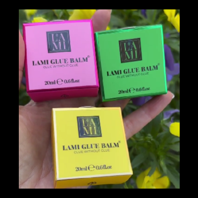 Caixas coloridas de LAMI GLUE BALM com texto preto seguradas à mão contra fundo de plantas com flores amarelas.