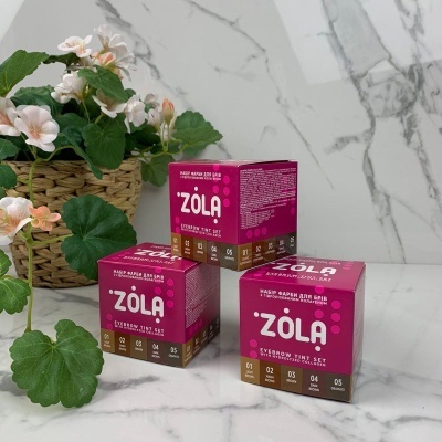 Três caixas coloridas do kit ZOLA para tintura de sobrancelhas ao lado de planta com flores