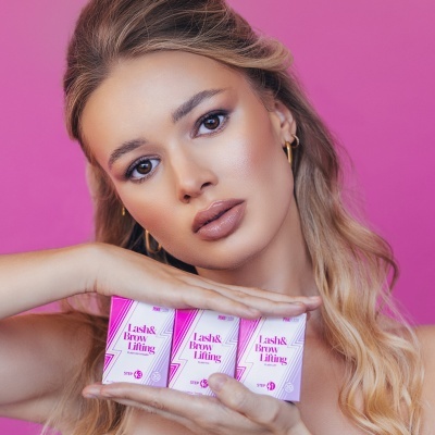 Mulher loira segurando caixas cor-de-rosa de Lash & Brow Lifting num fundo rosa