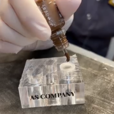 Frasco pequeno com líquido castanho e suporte transparente com texto AS COMPANY