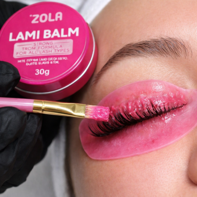 Pote rosa ZOLA LAMI BALM e pincel aplicando produto nas pestanas com patch rosa sob o olho.