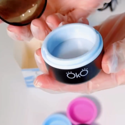 Frasco preto com tampa castanha aberto, pasta branca dentro, m 33os com luvas a segur e1-lo, tampas coloridas no fundo.