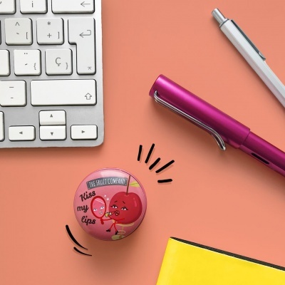 Latinha cor-de-rosa da YES FRUIT COMPANY com texto Kiss my lips junto a teclado, canetas e caderno sobre mesa laranja.