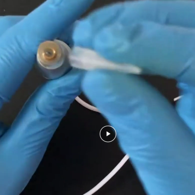 Peça metálica cilíndrica pequena com topo dourado sendo segurada por mãos com luvas azuis