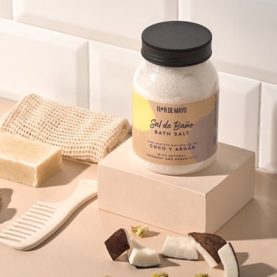 Frasco de sal de banho com aromas de coco e argan sobre caixa bege com pedaços de coco e acessórios de banho ao redor.