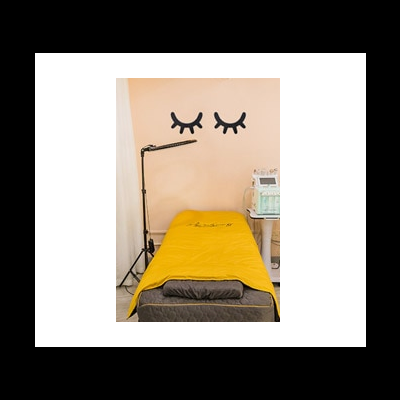 Cama de estética com colcha amarela e almofada cinza numa sala com parede bege decorada com pestanas negras