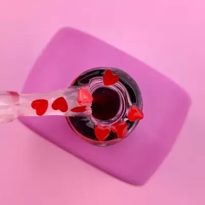 Frasco de vidro com gotas vermelhas em forma de coração sobre fundo rosa.