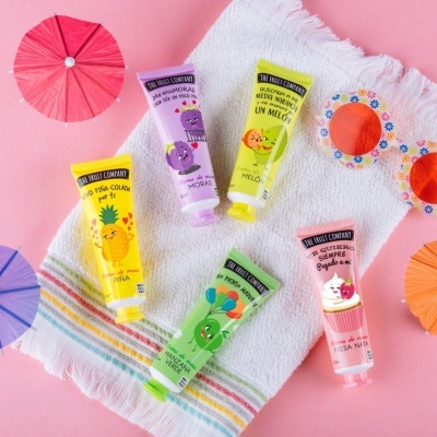 Cinco tubos coloridos da THE FRUIT COMPANY sobre toalha com guarda-sóis e óculos de sol em fundo rosa.