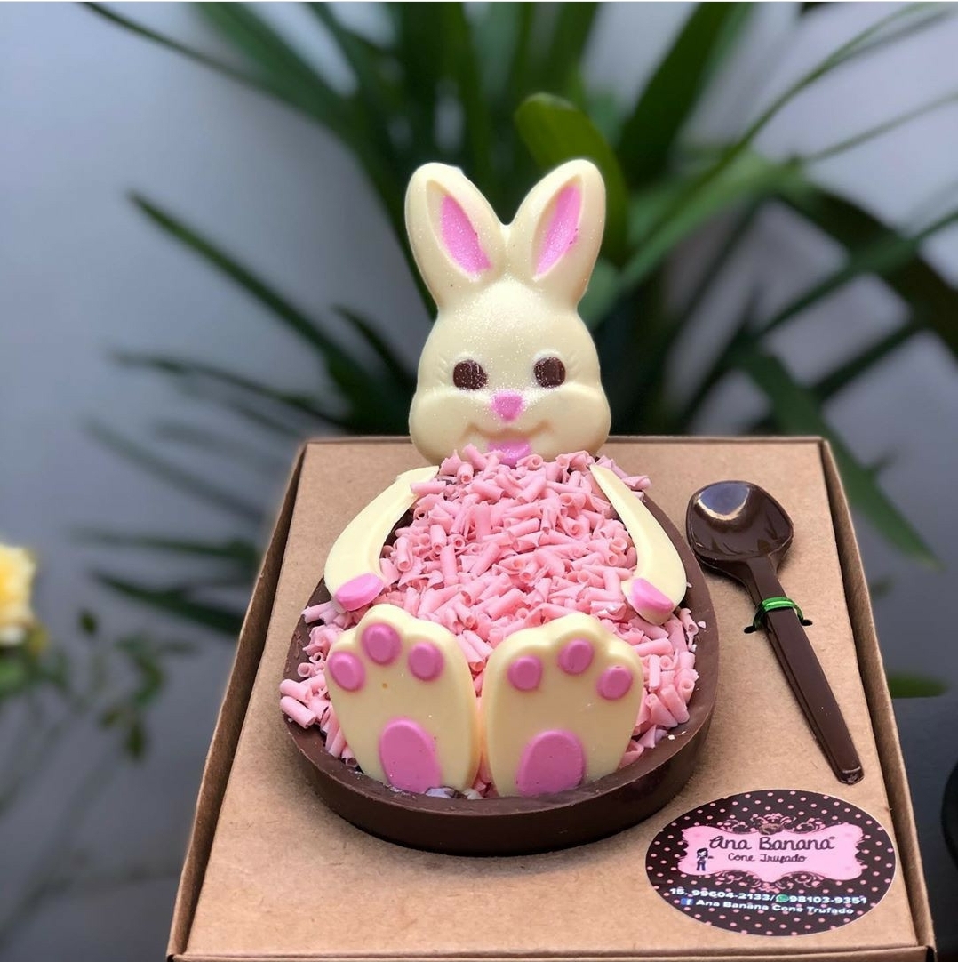 Ovo de chocolate com coelho de chocolate branco e confeitos rosa numa caixa de papelão com colher de chocolate