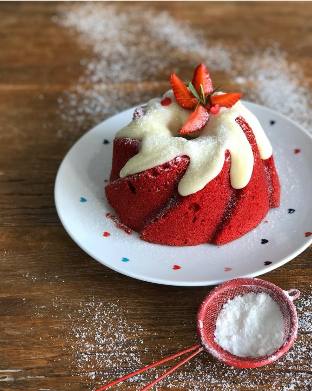 Bolo vermelho tulipa com calda branca e morangos em prato branco com corações, açúcar em coador vermelho sobre mesa de madeira.