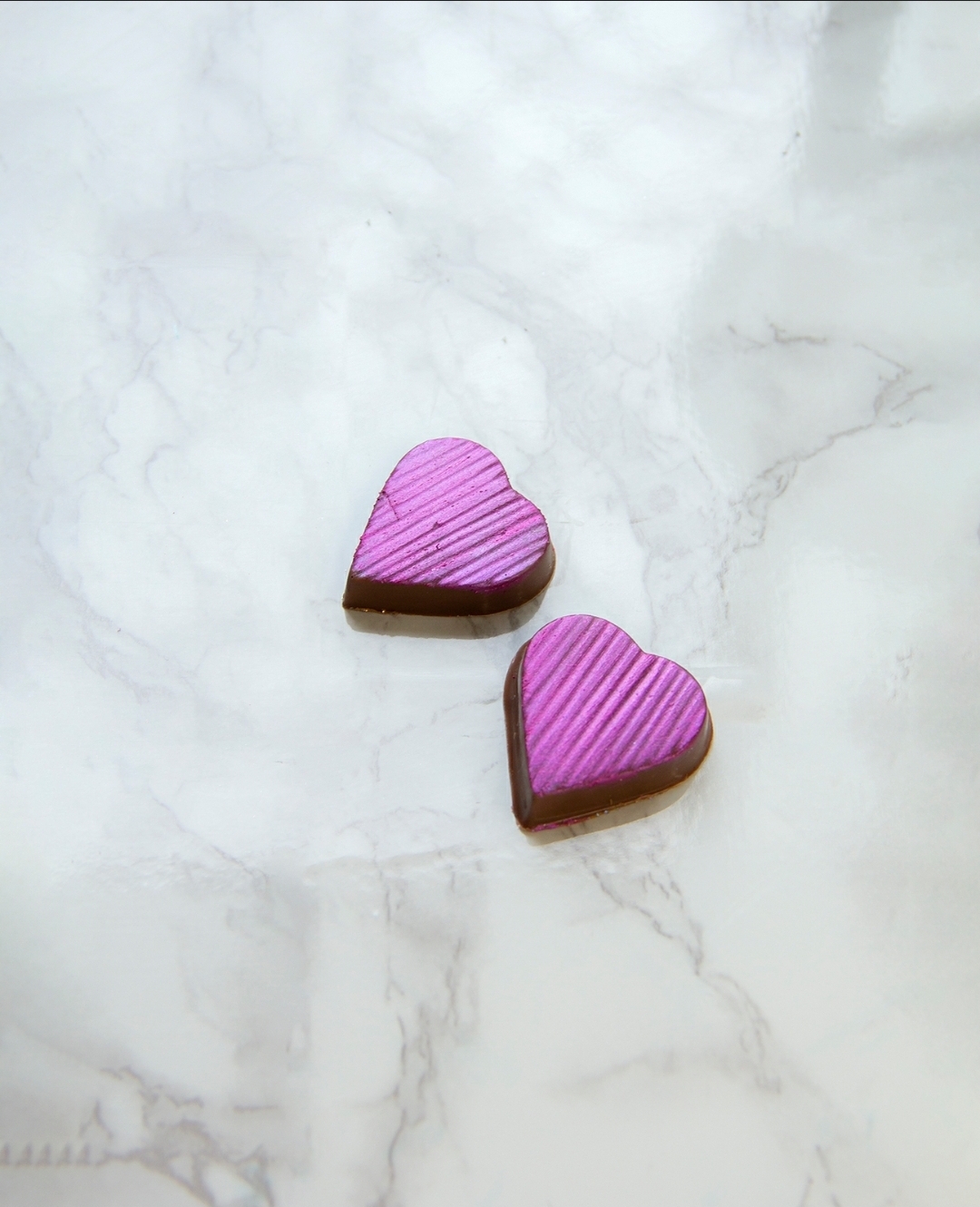 Dois chocolates em forma de coração roxos com textura riscada sobre fundo marmoreado branco.