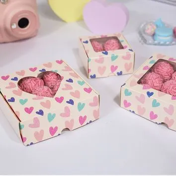 4331 Caixa Coração C/ Visor Amor em Cores- 9 doces-10 Unidades Caixas de papel com coração recortado, doces rosados dentro, superfície branca e fundo desfocado com itens coloridos.