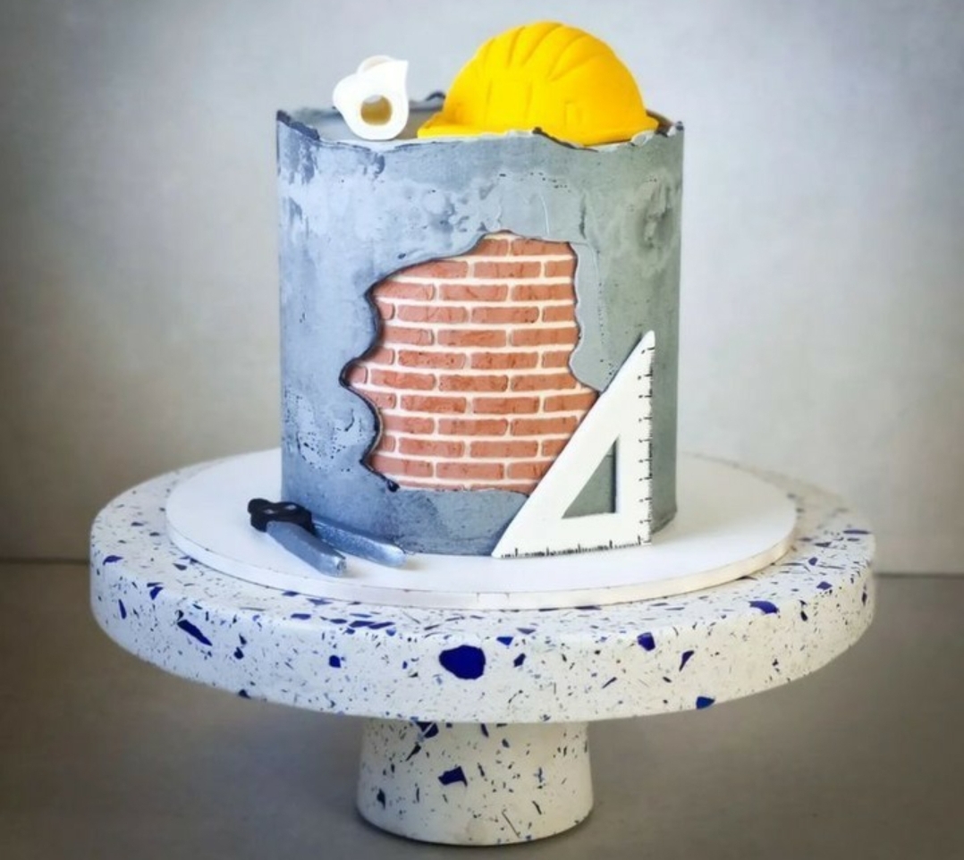 Bolo decorado com tema de construção com capacete amarelo e ferramentas em miniatura, sobre suporte branco com manchas azuis