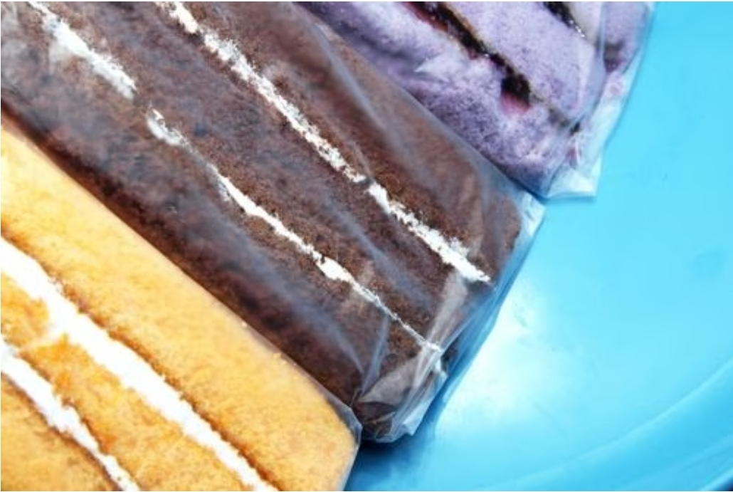 Três bolos fatiados de cores diferentes embalados em plástico sobre fundo azul