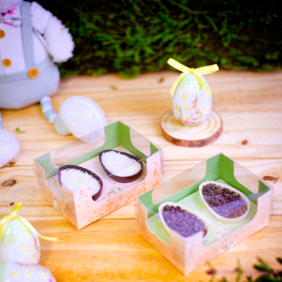 Caixas transparentes com ovos de chocolate decorados sobre mesa de madeira com decoração de coelhos e ovos coloridos