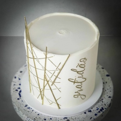 Bolo branco com decoração dourada e palavra gratidão em prato sobre base de padrão terrazzo