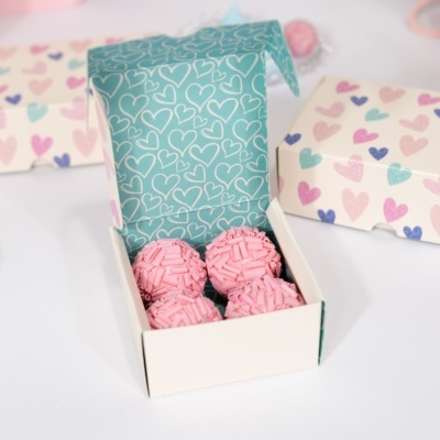 Caixa aberta com brigadeiros rosa e caixas decoradas com corações coloridos ao fundo.