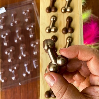 Mão segura chocolate dourado em forma de ossos, com mais chocolates semelhantes alinhados numa base de madeira e um molde de plástico ao lado.
