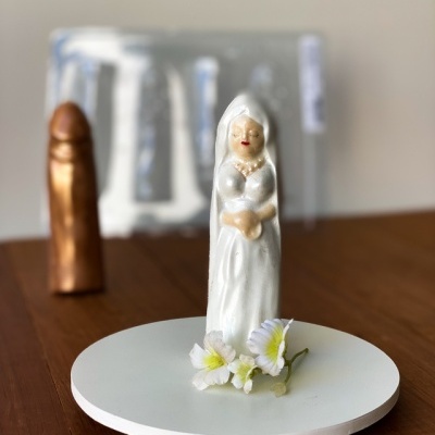Vela em forma de figura feminina branca sobre base redonda com flores, em mesa de madeira.