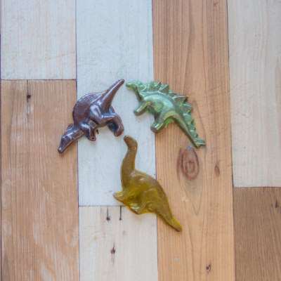 Ímanes de dinossauros castanho, verde metálico e amarelo em chão de madeira