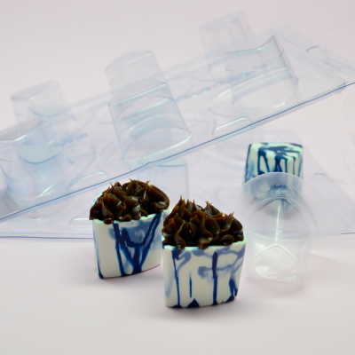 Dois chocolates decorados em azul e branco com cobertura de chocolate escuro ao lado de forma plástica transparente para moldar chocolate.