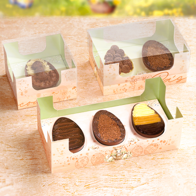 Três caixas transparentes com ovos de chocolate decorados em embalagem clara com motivos florais.