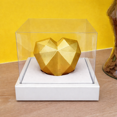 Coração geométrico dourado numa caixa transparente sobre base branca