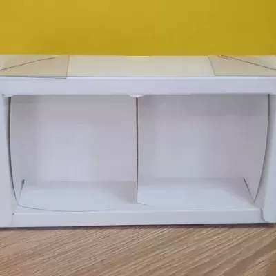 Caixa de embalagem branca aberta com duas divisórias, chão de madeira e parede amarela