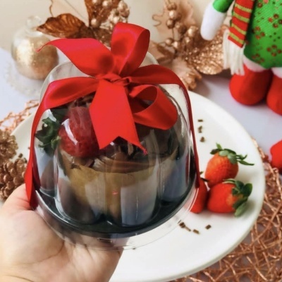 Pequeno bolo decorado com chocolate e morango dentro de embalagem plástica com laço vermelho.