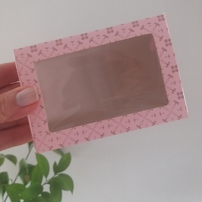 Caixa rosa com padrão de tesouras e janela transparente, mão segurando, planta ao fundo