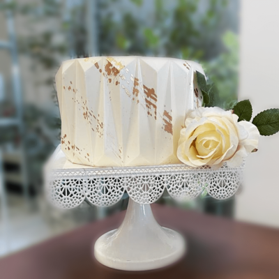 Bolo branco geométrico com detalhes dourados e rosa branca decorativa num suporte branco rendilhado