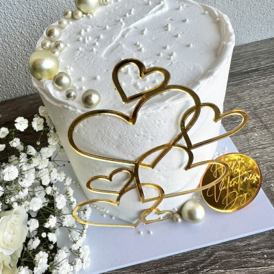 Bolo branco decorado com pérolas douradas e prateadas e enfeite dourado em forma de corações e círculo com texto em inglês