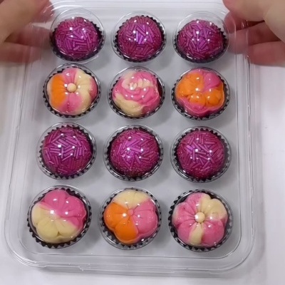 Doze doces decorativos coloridos em caixa transparente, com coberturas roxa e flores coloridas.