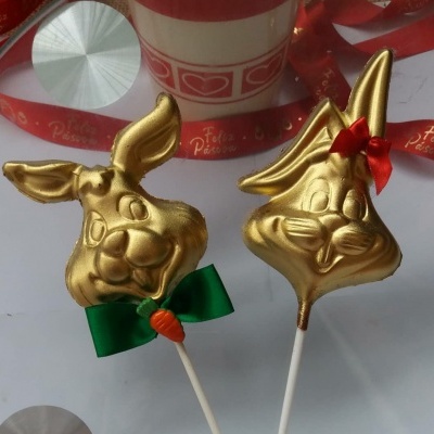 Dois pirulitos dourados em formato de coelho com laços e cenoura, mesa com fita 'Feliz Páscoa' e caneca.