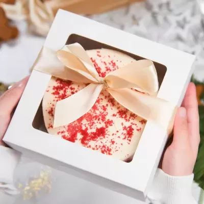 Bolo redondo com cobertura branca, pedaços vermelhos, caixa branca com laço creme