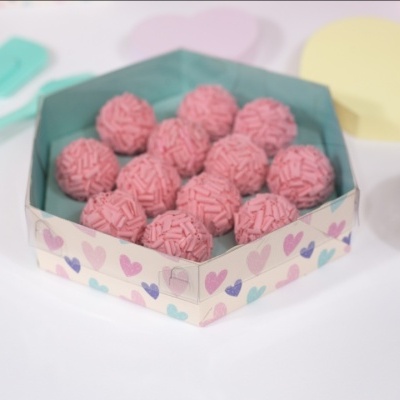 Caixa hexagonal com doces rosados cobertos por granulados em uma superfície branca com utensílios coloridos ao redor