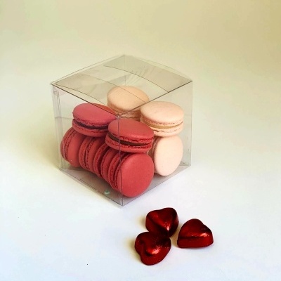 Caixa transparente com macarons vermelhos e bege junto a corações vermelhos metálicos.