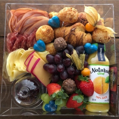 Caixa de tábuas de madeira com queijos, fiambre, chouriço, croissants, frutas, bombons, copo e sumo Natural One